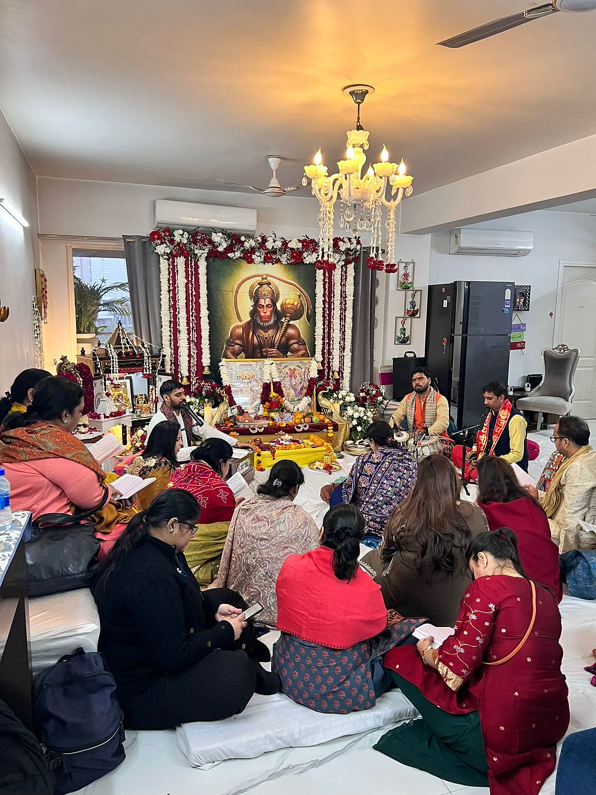 A full gathering seated for a Sunderkand Path at home in Delhi NCR, with the Hanuman ji mandap and the mandali