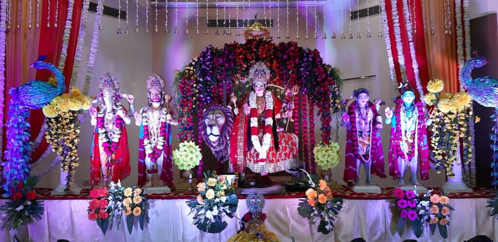 Mata Durga idol seated on her lion, flanked by Ganesh and Hanuman, decorated with flowers and peacock feathers for a Chowki darbar