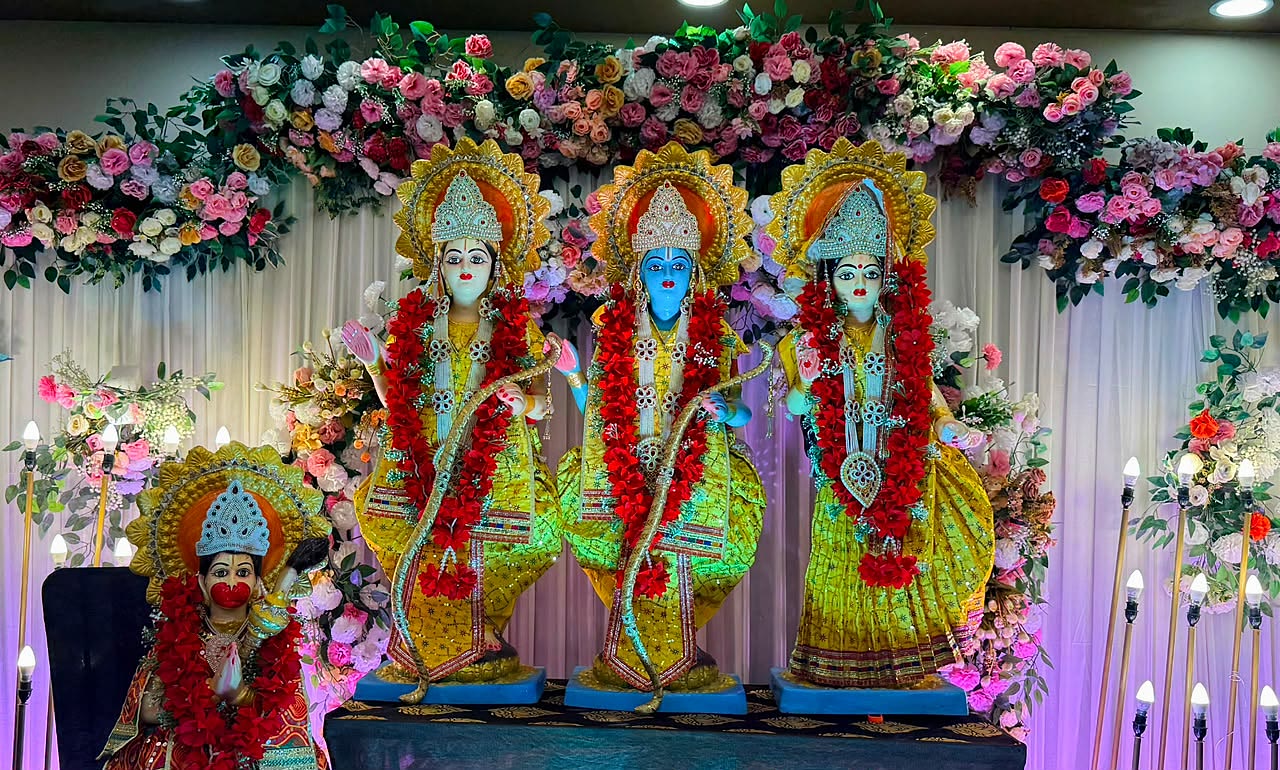 Wide shot of the Ram Darbar with Hanuman ji beneath a flower arch decorated with diyas